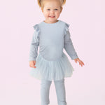 A smiling toddler with blond pigtails models the Isobella and Chloe "Tutu in the Clouds" Two Piece Pant Set—a light blue outfit with a ruffled top, tulle skirt, and matching leggings—standing against a light pink background.