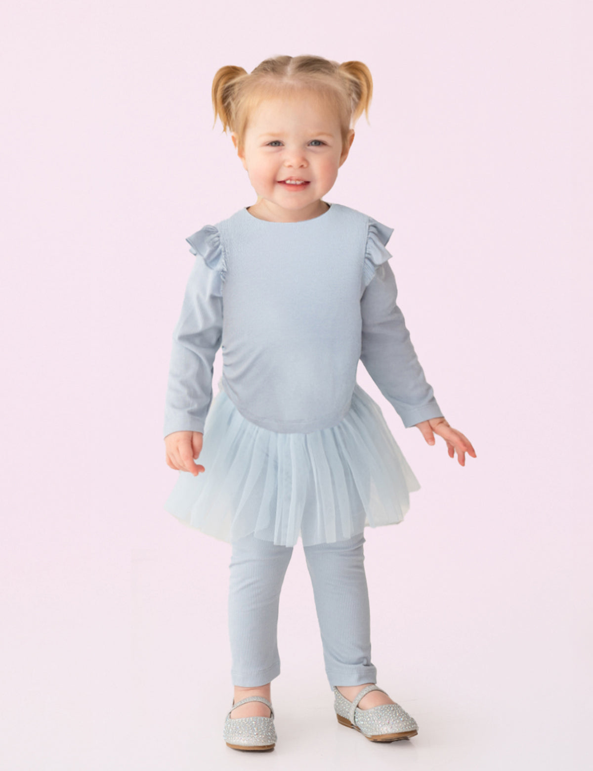 A smiling toddler with blond pigtails models the Isobella and Chloe "Tutu in the Clouds" Two Piece Pant Set—a light blue outfit with a ruffled top, tulle skirt, and matching leggings—standing against a light pink background.