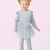 A smiling toddler with blond pigtails models the Isobella and Chloe "Tutu in the Clouds" Two Piece Pant Set—a light blue outfit with a ruffled top, tulle skirt, and matching leggings—standing against a light pink background.