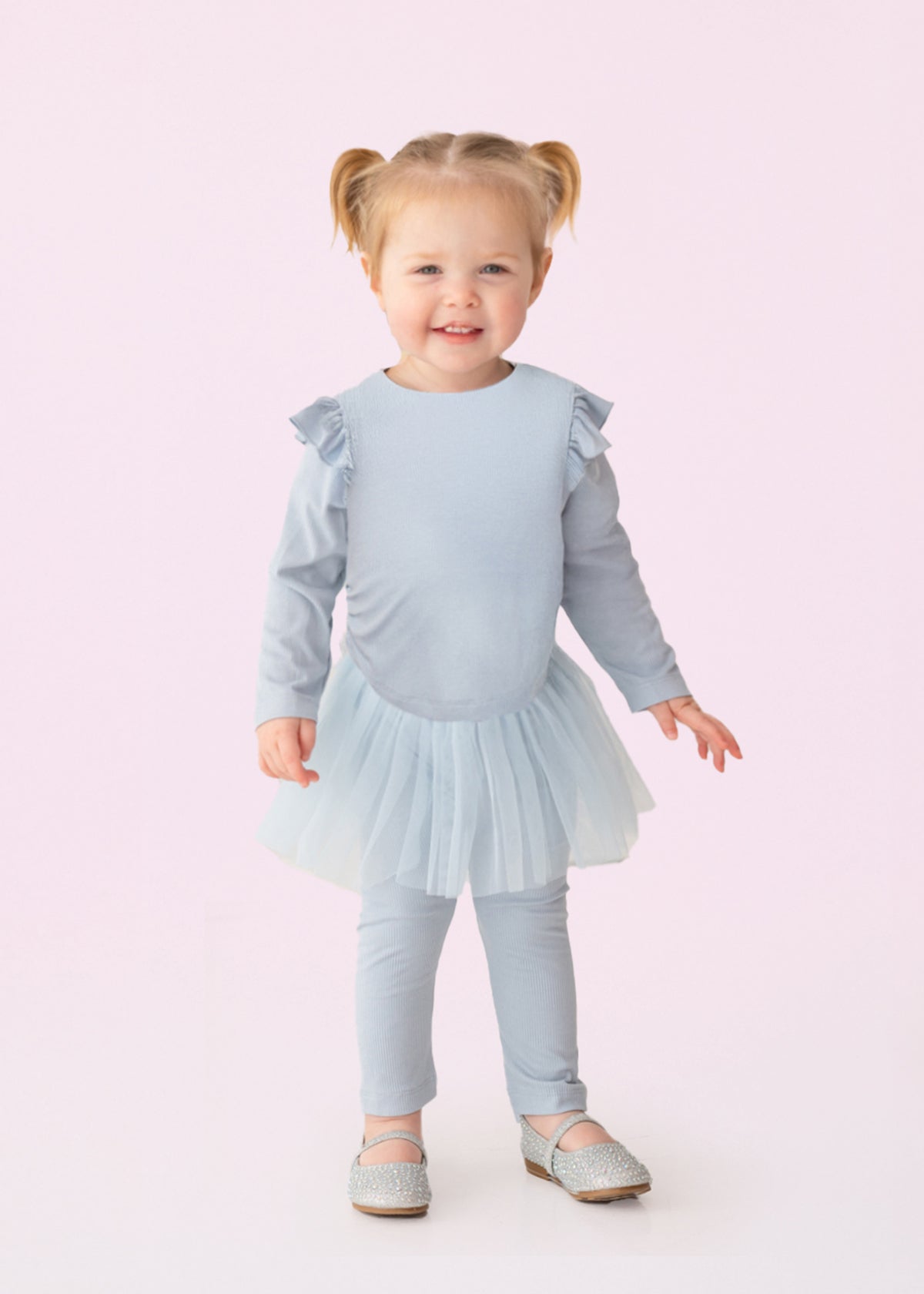 A smiling toddler with blond pigtails models the Isobella and Chloe "Tutu in the Clouds" Two Piece Pant Set—a light blue outfit with a ruffled top, tulle skirt, and matching leggings—standing against a light pink background.