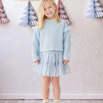 A young blonde girl wears the Isobella and Chloe "Tutu in the Clouds" two-piece set indoors on a wooden floor, with pastel paper Christmas trees hanging on strings against a textured white wall in the background.