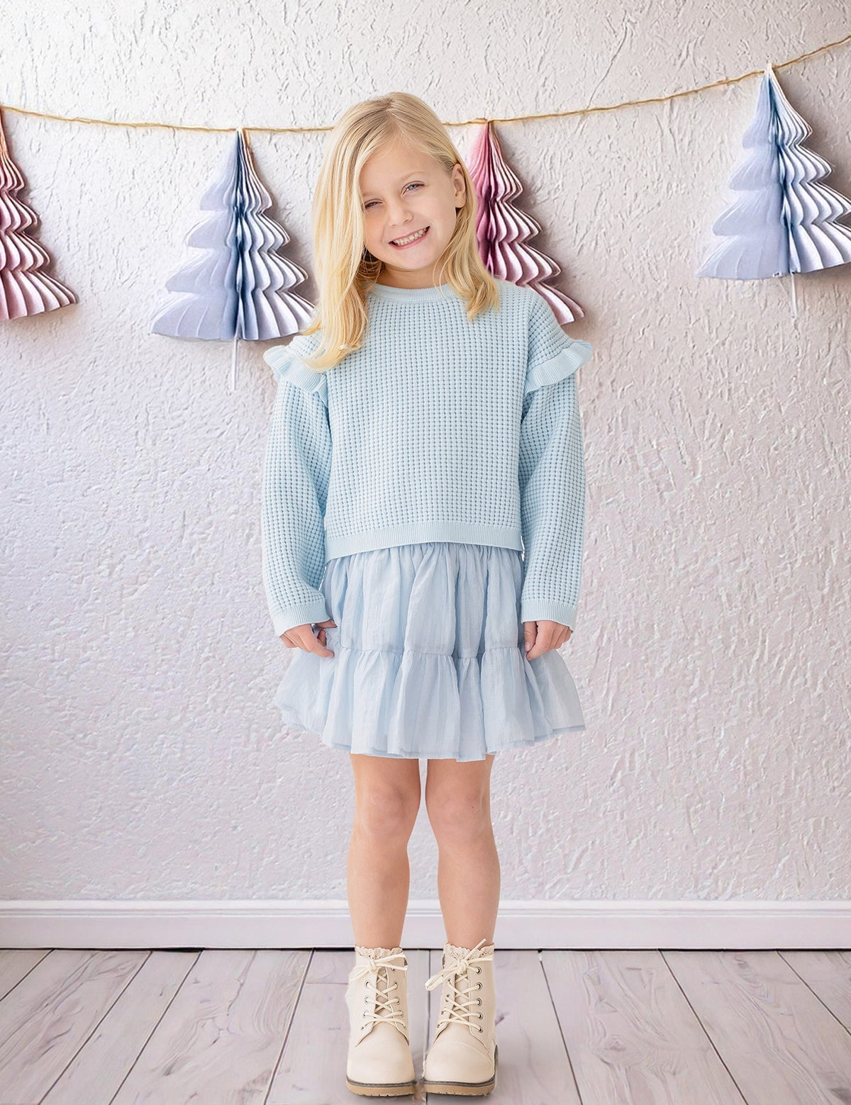 A young blonde girl wears the Isobella and Chloe "Tutu in the Clouds" two-piece set indoors on a wooden floor, with pastel paper Christmas trees hanging on strings against a textured white wall in the background.
