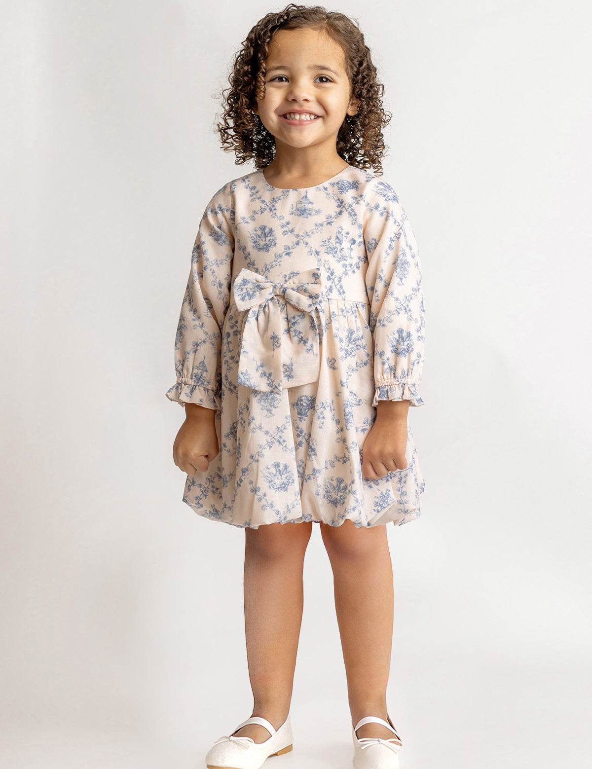 A young girl with curly hair smiles in a Mabel and Honey Princess Dress, featuring blue floral fabric and a waist bow, paired with white dress shoes against a plain light background.