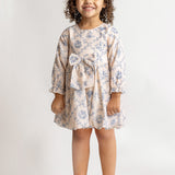 A young girl with curly hair smiles in a Mabel and Honey Princess Dress, featuring blue floral fabric and a waist bow, paired with white dress shoes against a plain light background.