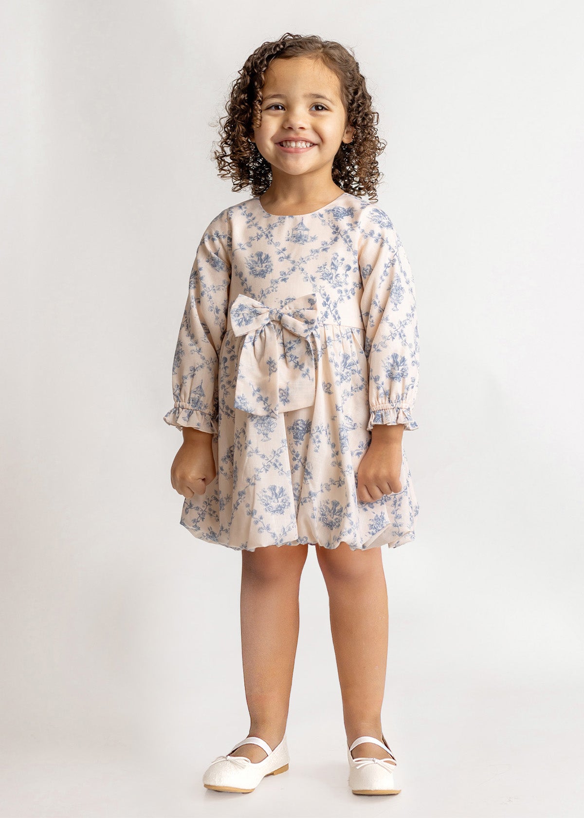 A young girl with curly hair smiles in a Mabel and Honey Princess Dress, featuring blue floral fabric and a waist bow, paired with white dress shoes against a plain light background.