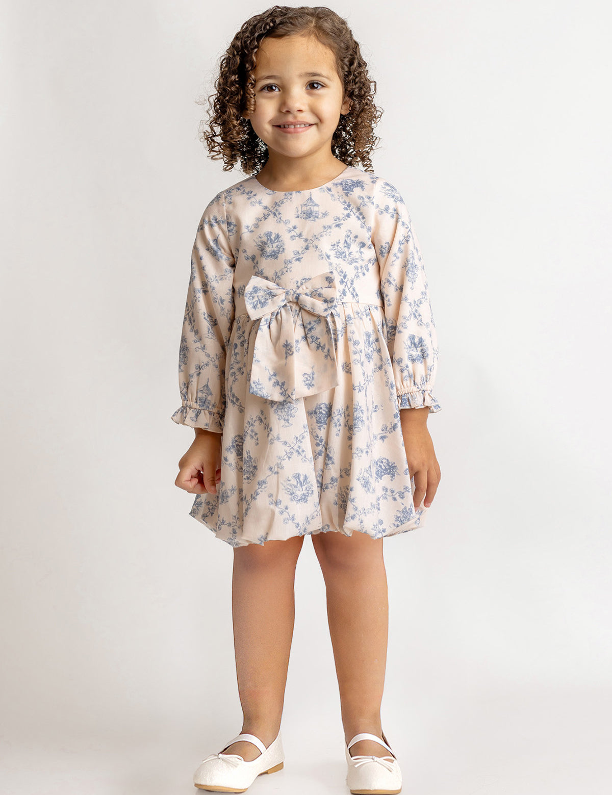 A young girl with curly hair smiles against a plain background, wearing the Mabel and Honey Princess Dress—a blue floral long-sleeve dress with a bow—paired with white shoes for a charming princess look.