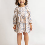 A young girl with curly hair smiles against a plain background, wearing the Mabel and Honey Princess Dress—a blue floral long-sleeve dress with a bow—paired with white shoes for a charming princess look.