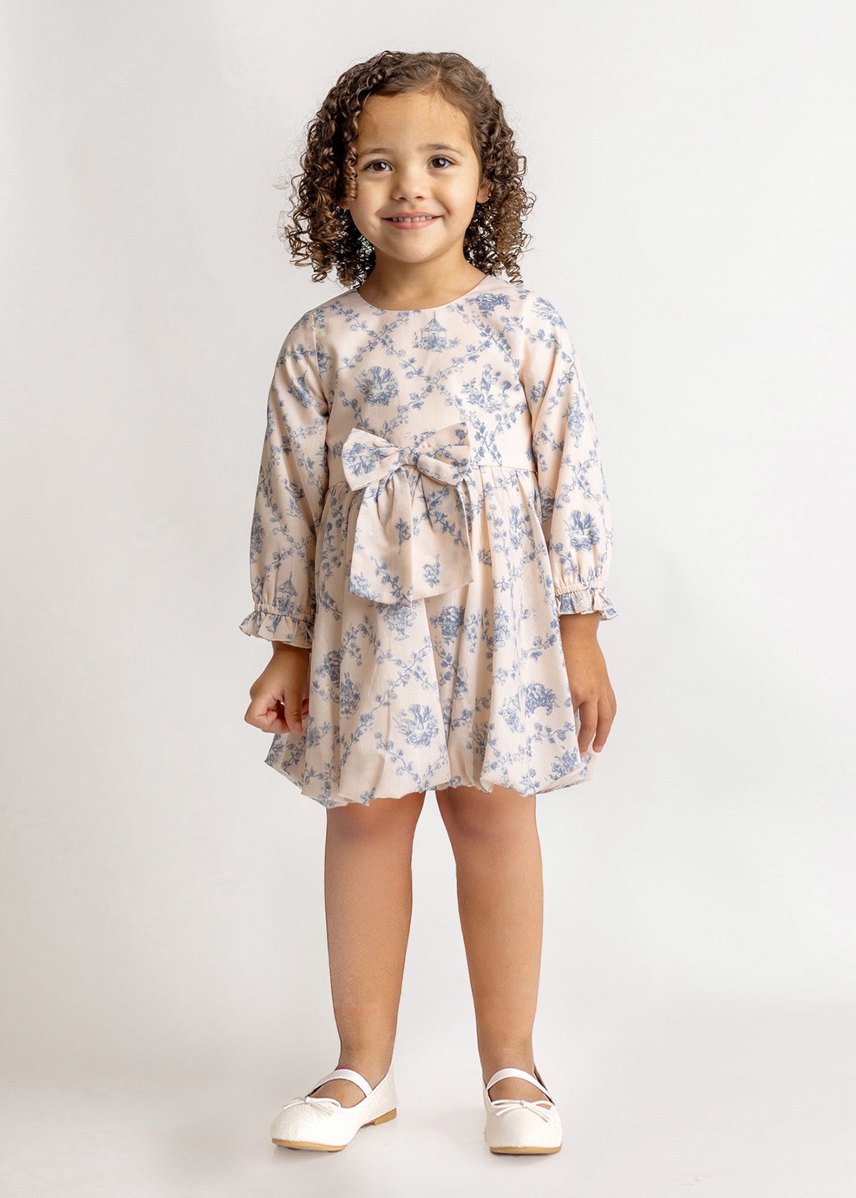 A young girl with curly hair smiles against a plain background, wearing the Mabel and Honey Princess Dress—a blue floral long-sleeve dress with a bow—paired with white shoes for a charming princess look.