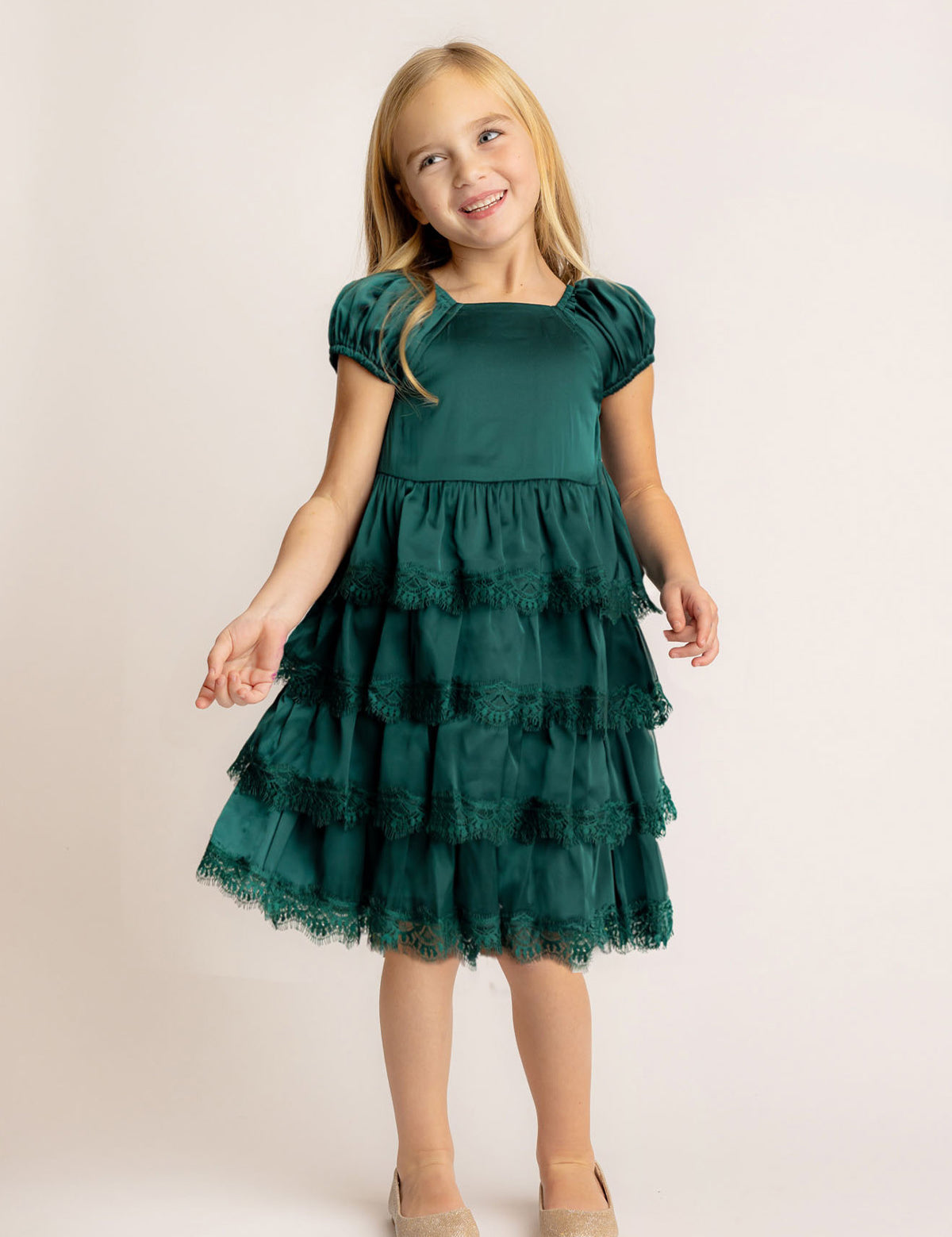 A young girl with long blonde hair smiles while wearing the Isobella and Chloe Mistletoe Emerald Green Tiered Dress with puffed sleeves and lace trim, paired with beige shoes, standing against a plain light background.