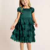 A young girl with long blonde hair smiles while wearing the Isobella and Chloe Mistletoe Emerald Green Tiered Dress with puffed sleeves and lace trim, paired with beige shoes, standing against a plain light background.