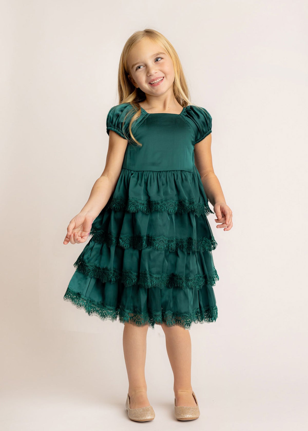 A young girl with long blonde hair smiles while wearing the Isobella and Chloe Mistletoe Emerald Green Tiered Dress with puffed sleeves and lace trim, paired with beige shoes, standing against a plain light background.