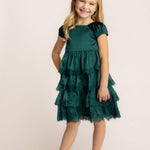 A young girl with long blonde hair smiles in front of a light background, wearing the Isobella and Chloe Mistletoe Emerald Green Tiered Dress with lace trim and sparkly beige ballet flats.
