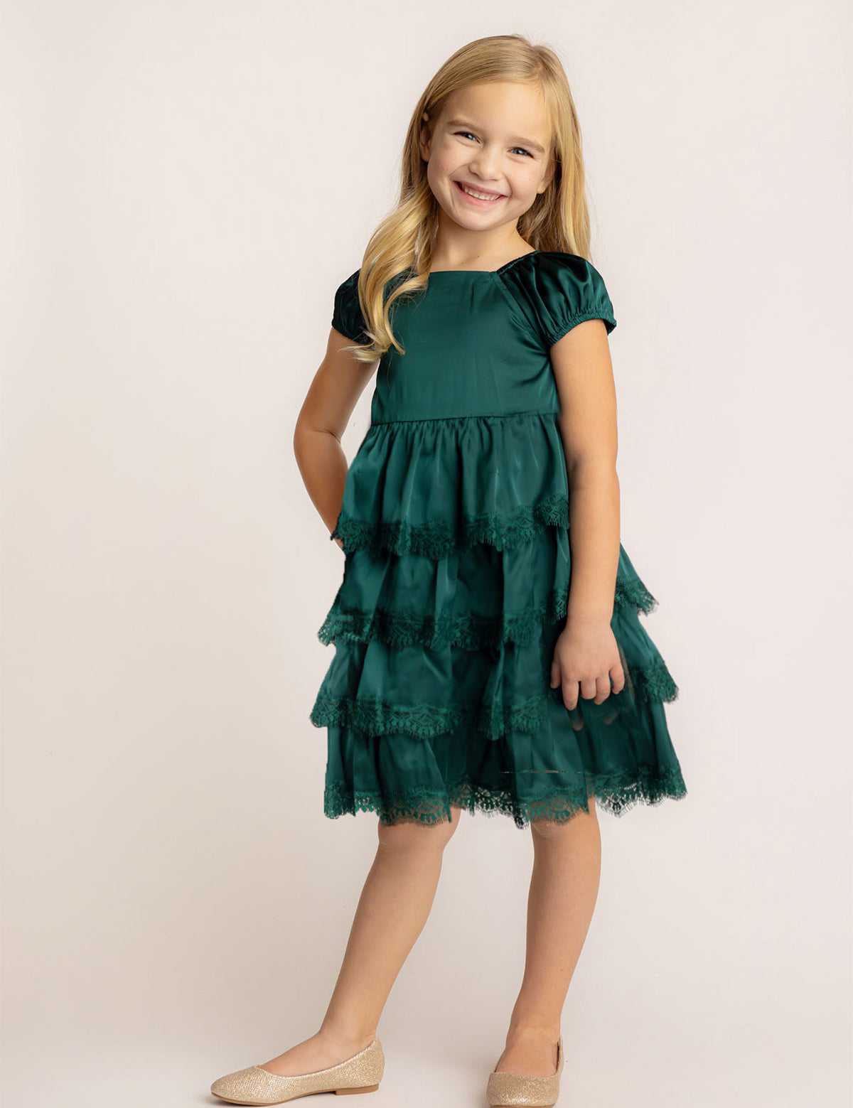 A young girl with long blonde hair smiles in front of a light background, wearing the Isobella and Chloe Mistletoe Emerald Green Tiered Dress with lace trim and sparkly beige ballet flats.