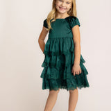 A young girl with long blonde hair smiles in front of a light background, wearing the Isobella and Chloe Mistletoe Emerald Green Tiered Dress with lace trim and sparkly beige ballet flats.