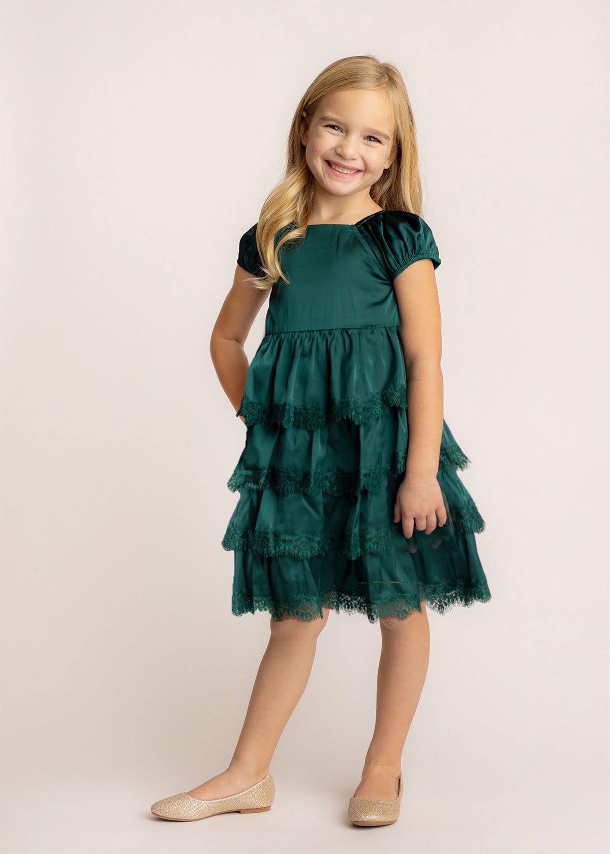A young girl with long blonde hair smiles in front of a light background, wearing the Isobella and Chloe Mistletoe Emerald Green Tiered Dress with lace trim and sparkly beige ballet flats.