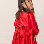 A young girl in the Isobella and Chloe Ruby Red Tiered Dress, featuring a large back bow, stands facing away with a smile over her shoulder. She has long dark hair, and the background is plain and light-colored.