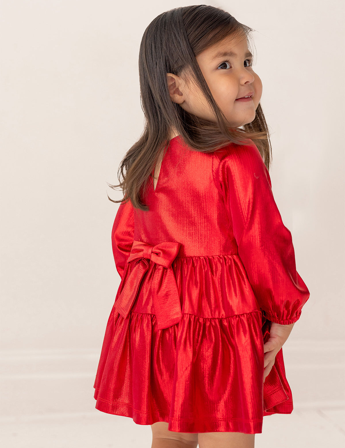 A young girl in the Isobella and Chloe Ruby Red Tiered Dress, featuring a large back bow, stands facing away with a smile over her shoulder. She has long dark hair, and the background is plain and light-colored.