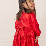 A young girl in the Isobella and Chloe Ruby Red Tiered Dress, featuring a large back bow, stands facing away with a smile over her shoulder. She has long dark hair, and the background is plain and light-colored.