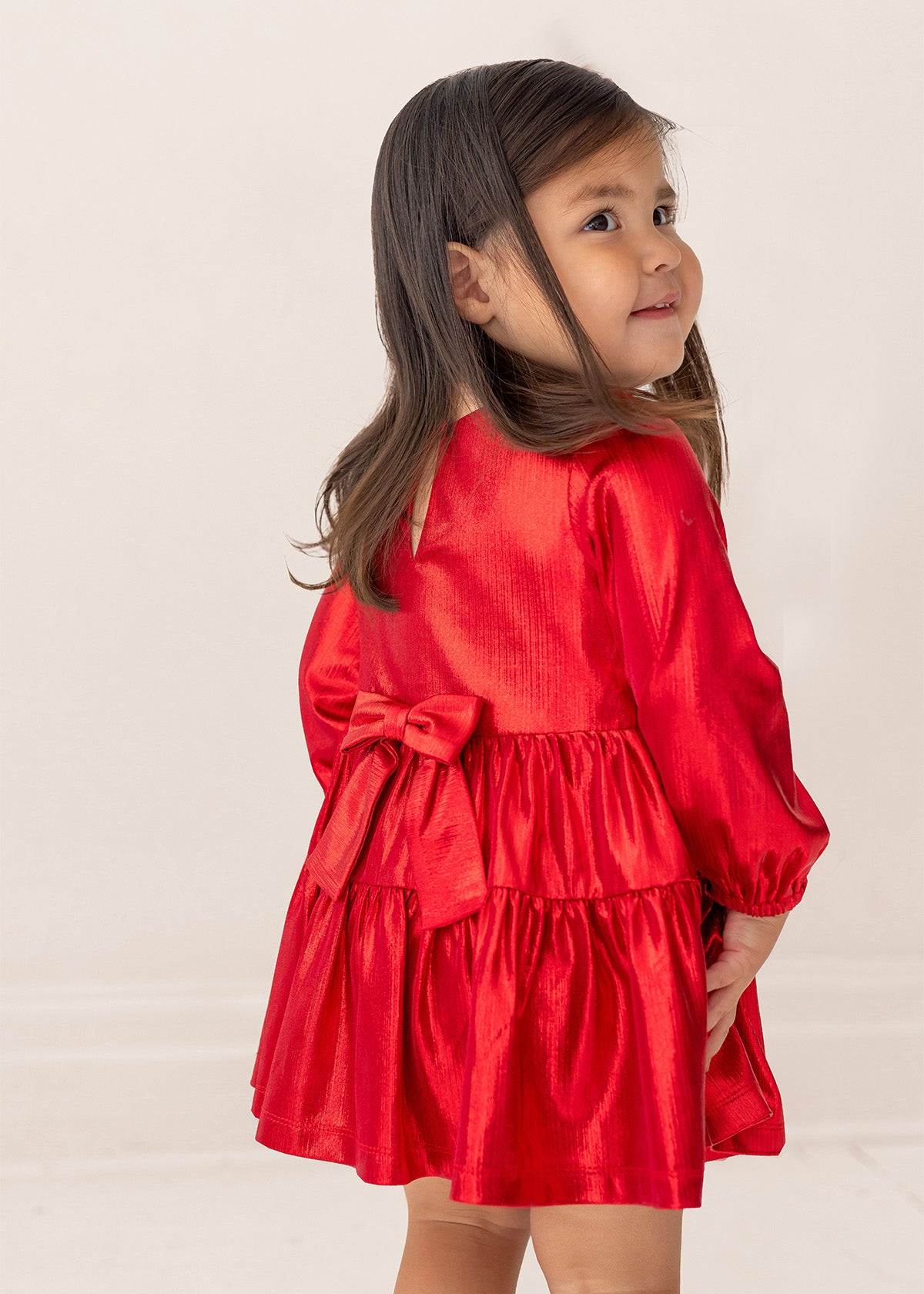 A young girl in the Isobella and Chloe Ruby Red Tiered Dress, featuring a large back bow, stands facing away with a smile over her shoulder. She has long dark hair, and the background is plain and light-colored.