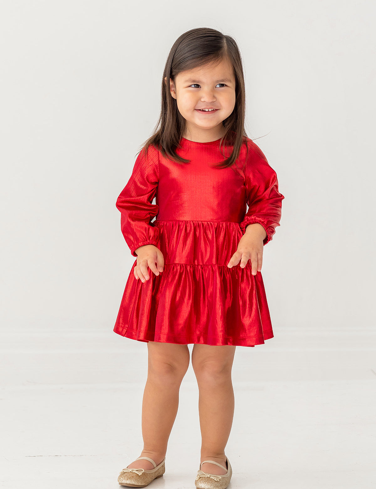 A young girl with long brown hair smiles in the Isobella and Chloe Ruby Red Teired Dress, featuring long sleeves and a ruffled skirt, paired with gold shoes, standing against a plain white background.
