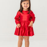 A young girl with long brown hair smiles in the Isobella and Chloe Ruby Red Teired Dress, featuring long sleeves and a ruffled skirt, paired with gold shoes, standing against a plain white background.