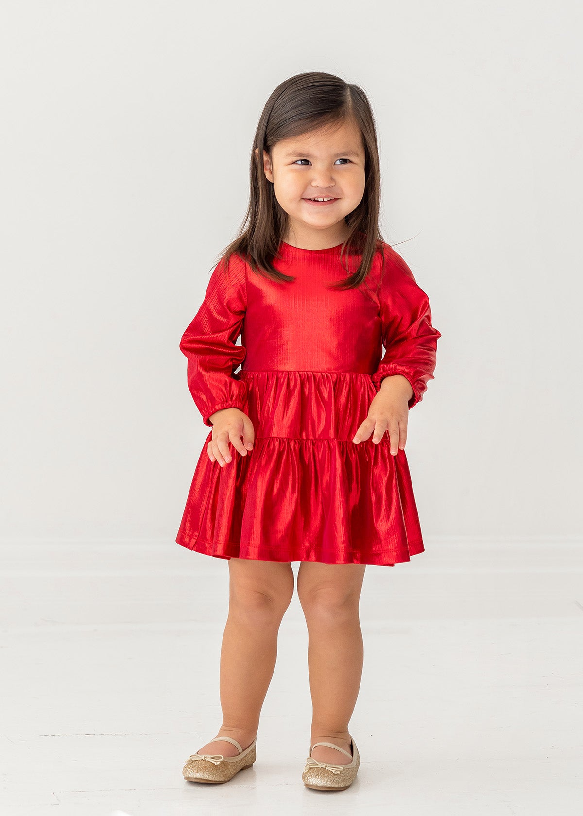 A young girl with long brown hair smiles in the Isobella and Chloe Ruby Red Teired Dress, featuring long sleeves and a ruffled skirt, paired with gold shoes, standing against a plain white background.