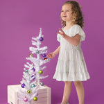 A young child wears the Sparkle and Spin Silver Dress by Isobella and Chloe, standing beside a small white Christmas tree with colorful ornaments, set on a wooden crate against a purple background.