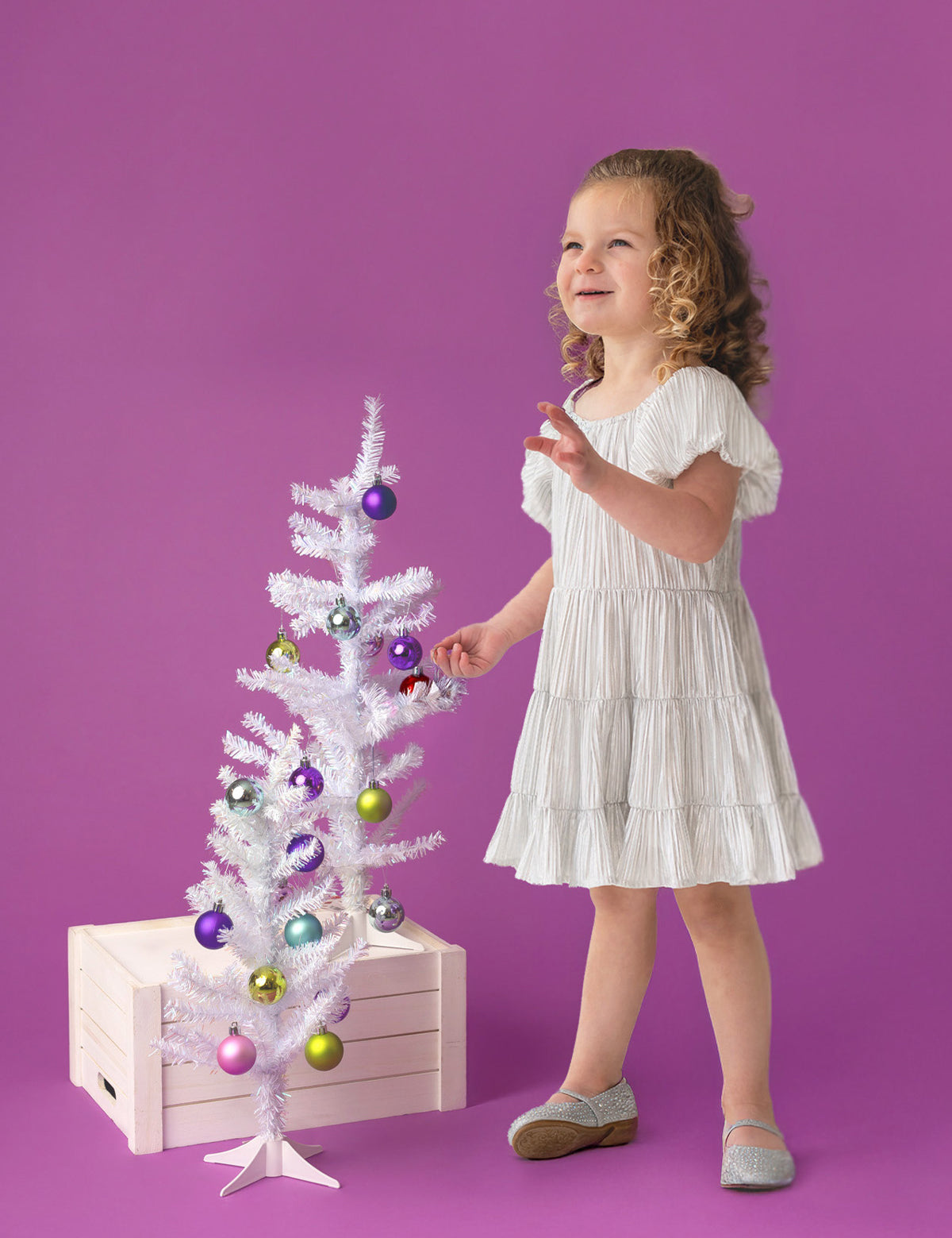 A young child wears the Sparkle and Spin Silver Dress by Isobella and Chloe, standing beside a small white Christmas tree with colorful ornaments, set on a wooden crate against a purple background.