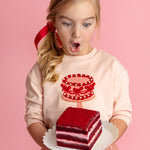 A young blond girl with a red ribbon, wearing the Isobella and Chloe Sweet Cakes Two Piece Set, looks surprised and excited as she holds a plate with red velvet cake against a pink background.