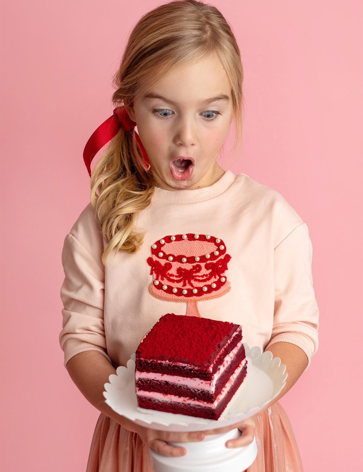 A young blond girl with a red ribbon, wearing the Isobella and Chloe Sweet Cakes Two Piece Set, looks surprised and excited as she holds a plate with red velvet cake against a pink background.