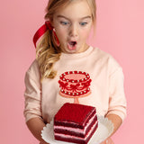 A young blond girl with a red ribbon, wearing the Isobella and Chloe Sweet Cakes Two Piece Set, looks surprised and excited as she holds a plate with red velvet cake against a pink background.