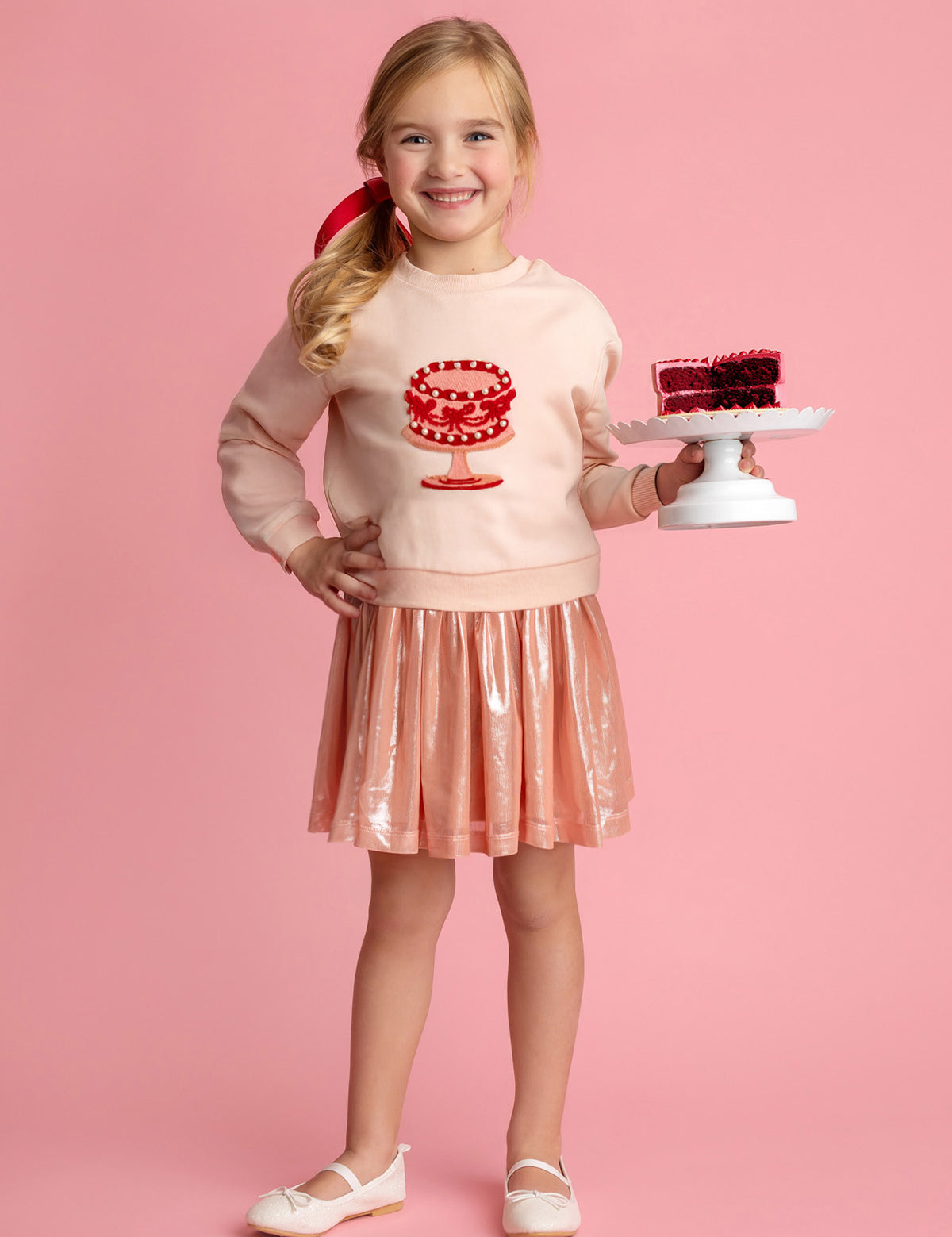 A smiling young girl stands against a pink background, wearing the Isobella and Chloe Sweet Cakes Two Piece Set, featuring an embroidered cake sweater and shiny pink skirt, while holding a small cake with red decoration on a white stand.