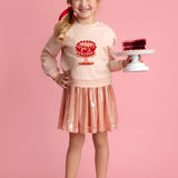 A smiling young girl stands against a pink background, wearing the Isobella and Chloe Sweet Cakes Two Piece Set, featuring an embroidered cake sweater and shiny pink skirt, while holding a small cake with red decoration on a white stand.