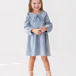 A young girl with light brown hair smiles while standing against a white background, wearing the Mabel and Honey Denim Days Long Sleeve Dress styled with beige ankle boots.