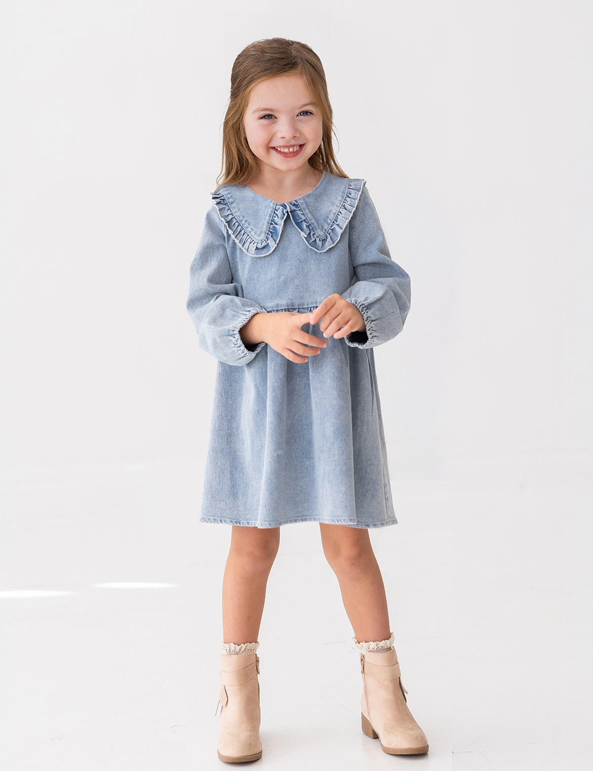 A young girl with light brown hair smiles while standing against a white background, wearing the Mabel and Honey Denim Days Long Sleeve Dress styled with beige ankle boots.