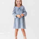 A young girl with light brown hair smiles while standing against a white background, wearing the Mabel and Honey Denim Days Long Sleeve Dress styled with beige ankle boots.