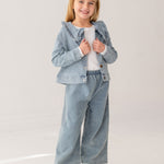 A young girl with blonde hair smiles in the Mabel and Honey Denim Days Ruffled Collar Jacket, paired with matching wide-leg denim pants, a white shirt, and white sneakers against a plain light background.