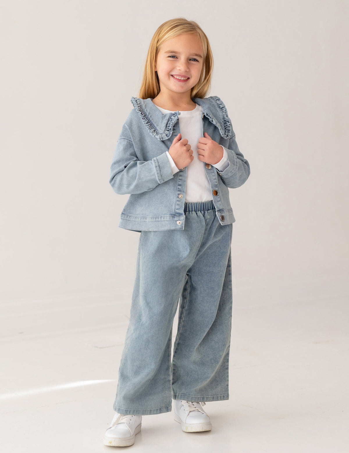 A young girl with blonde hair smiles in the Mabel and Honey Denim Days Ruffled Collar Jacket, paired with matching wide-leg denim pants, a white shirt, and white sneakers against a plain light background.