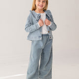 A young girl with blonde hair smiles in the Mabel and Honey Denim Days Ruffled Collar Jacket, paired with matching wide-leg denim pants, a white shirt, and white sneakers against a plain light background.