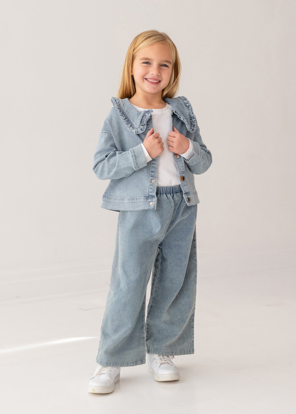 A young girl with blonde hair smiles in the Mabel and Honey Denim Days Ruffled Collar Jacket, paired with matching wide-leg denim pants, a white shirt, and white sneakers against a plain light background.
