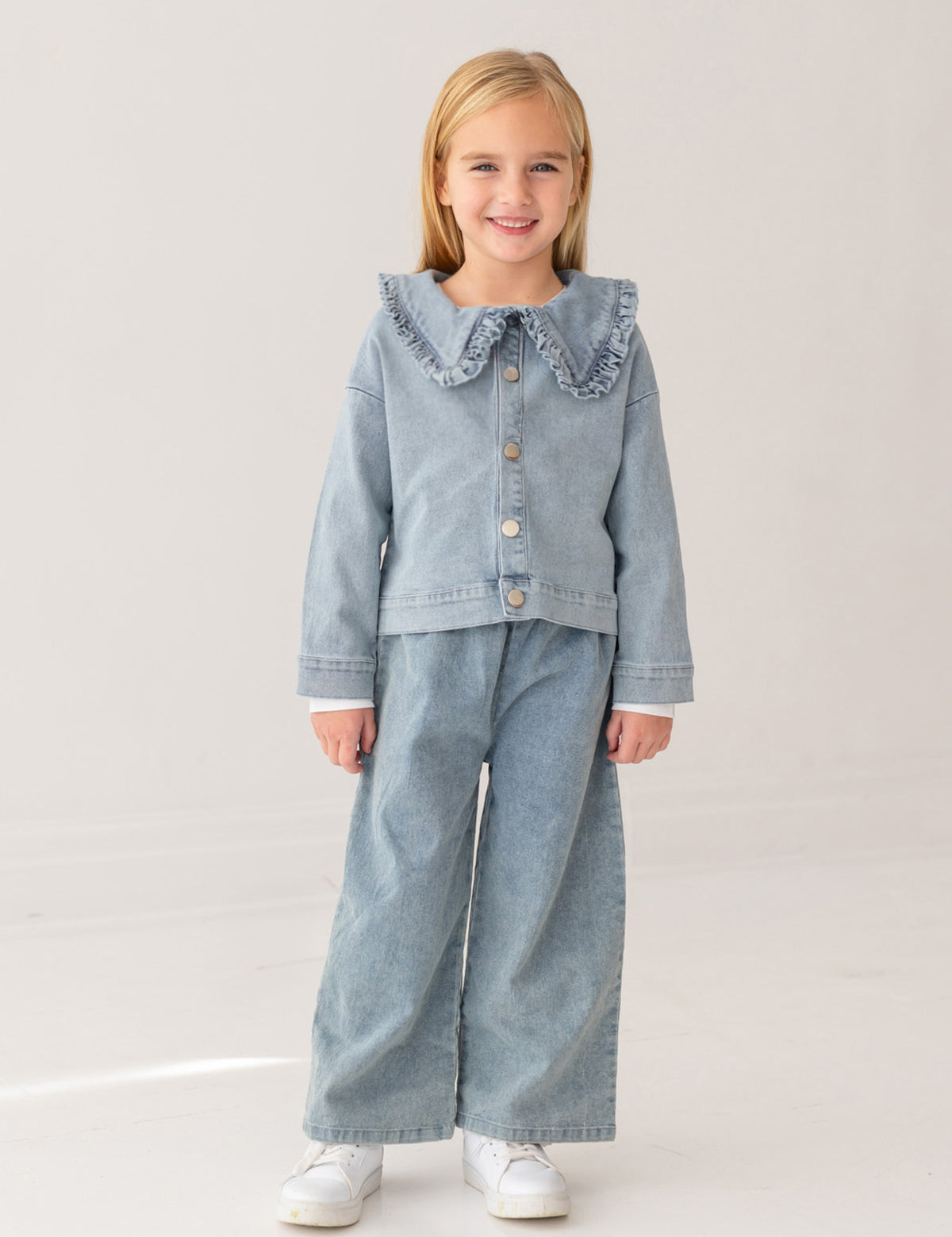 A young blonde girl smiles in a Mabel and Honey Denim Days Ruffled Collar Jacket, paired with matching wide-leg denim pants and white sneakers, standing against a plain light background—a stylish kids’ wardrobe choice.