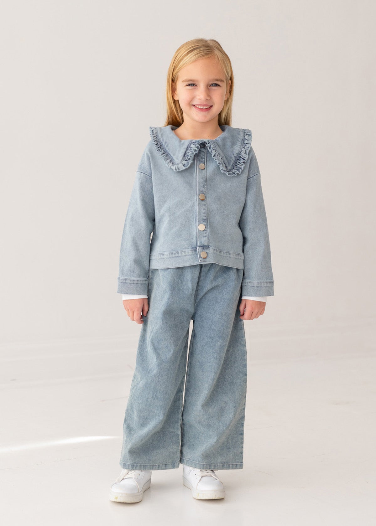 A young blonde girl smiles in a Mabel and Honey Denim Days Ruffled Collar Jacket, paired with matching wide-leg denim pants and white sneakers, standing against a plain light background—a stylish kids’ wardrobe choice.