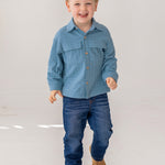 A young boy with short light brown hair smiles as he walks, wearing the Beckett and Bear Lucas Jacket over a light blue shirt, dark blue jeans, and brown shoes against a plain white background.