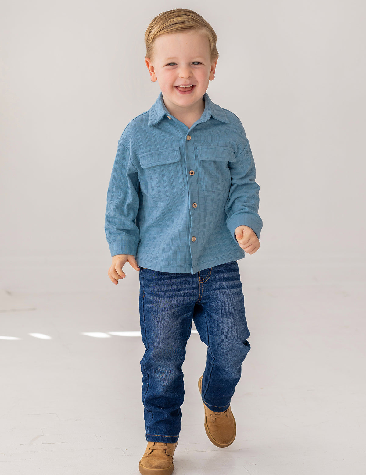 A young boy with short light brown hair smiles as he walks, wearing the Beckett and Bear Lucas Jacket over a light blue shirt, dark blue jeans, and brown shoes against a plain white background.