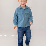 A young boy with short light brown hair smiles as he walks, wearing the Beckett and Bear Lucas Jacket over a light blue shirt, dark blue jeans, and brown shoes against a plain white background.