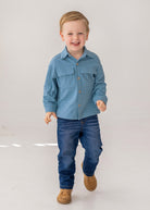 A young boy with short light brown hair smiles as he walks, wearing the Beckett and Bear Lucas Jacket over a light blue shirt, dark blue jeans, and brown shoes against a plain white background.