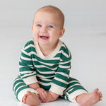 A smiling baby sits barefoot on a light floor, wearing Beckett and Bear's Jackson Romper—a green and white striped long-sleeve outfit—against a plain background.