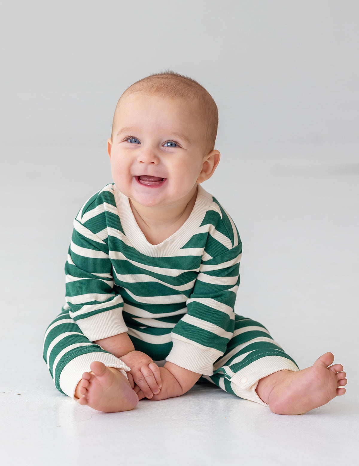 A smiling baby sits barefoot on a light floor, wearing Beckett and Bear's Jackson Romper—a green and white striped long-sleeve outfit—against a plain background.