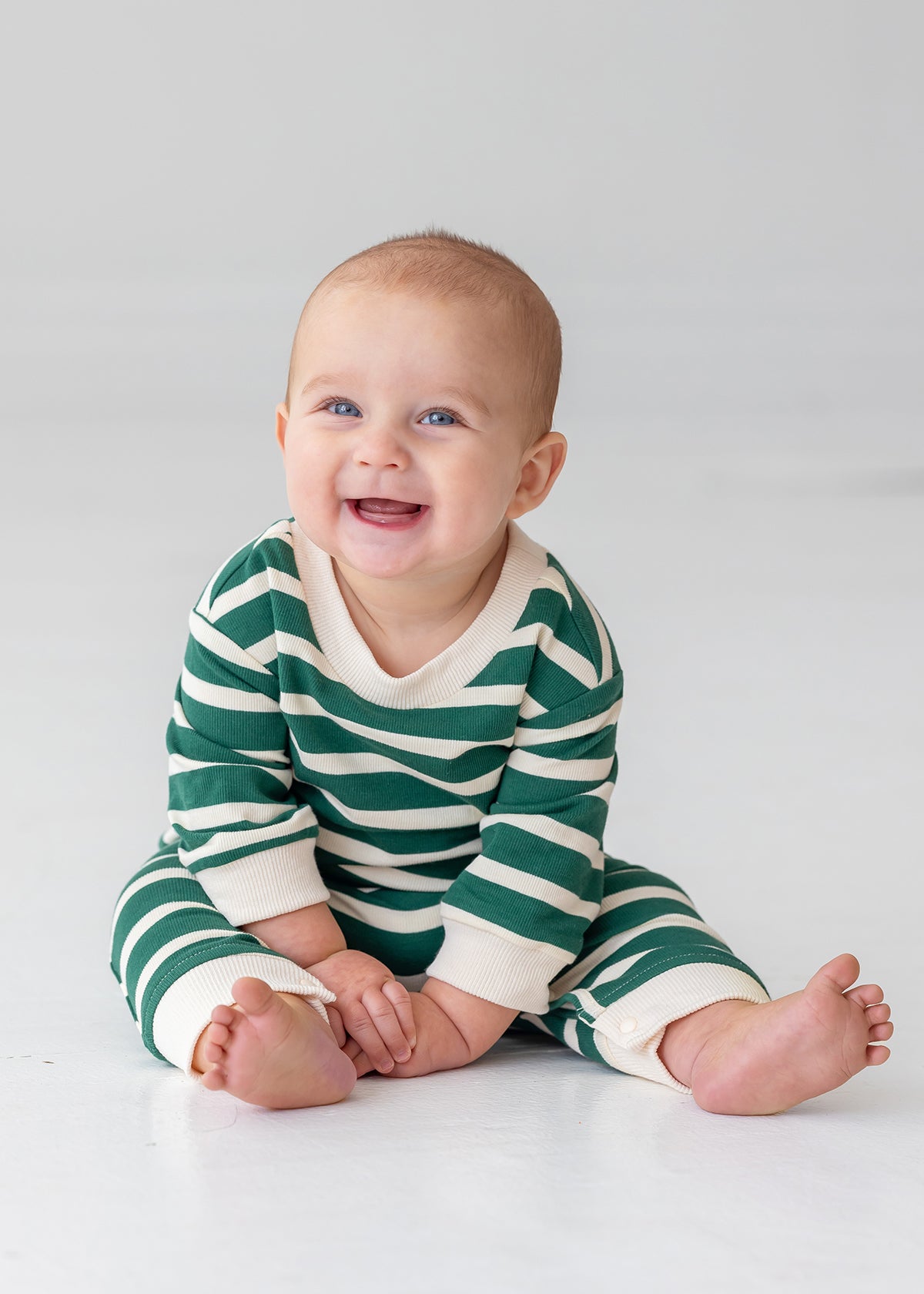 A smiling baby sits barefoot on a light floor, wearing Beckett and Bear's Jackson Romper—a green and white striped long-sleeve outfit—against a plain background.