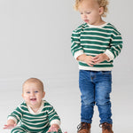 A baby smiles brightly while sitting barefoot in the Beckett and Bear Jackson Romper, next to a toddler in jeans and brown boots. Both are on a white floor, dressed in green and white stripes.